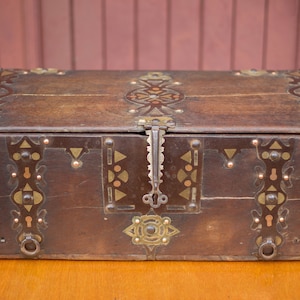 May include: A wooden chest with intricate brass and copper accents. The chest has a hinged lid and a lock with a keyhole. The chest is decorated with geometric patterns and is a beautiful example of craftsmanship.