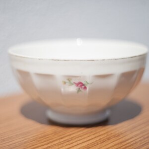 K&ouml;nnte beinhalten: Eine wei&szlig;e Keramiksch&uuml;ssel mit geriffeltem Design und einem Blumenmuster. Die Sch&uuml;ssel hat eine kleine rosa Rose mit gr&uuml;nen Bl&auml;ttern.