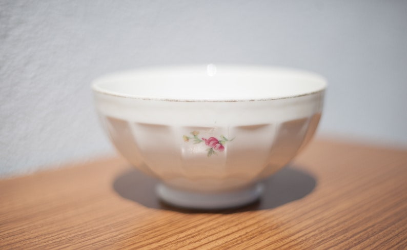 K&ouml;nnte beinhalten: Eine wei&szlig;e Keramiksch&uuml;ssel mit geriffeltem Design und einem Blumenmuster. Die Sch&uuml;ssel hat eine kleine rosa Rose mit gr&uuml;nen Bl&auml;ttern.