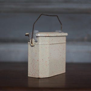 Peut inclure: Petite boîte rectangulaire en métal blanc cassé, avec un couvercle à charnière et une poignée en fil de fer. Le récipient présente un motif tacheté de points rouges, probablement vintage.