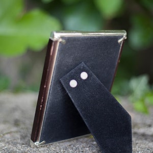 May include: A small, rectangular picture frame with a dark brown border and a silver-colored trim. The back of the frame is black, with a black velvet easel stand. The frame is set on a gray stone surface, with green foliage in the background.