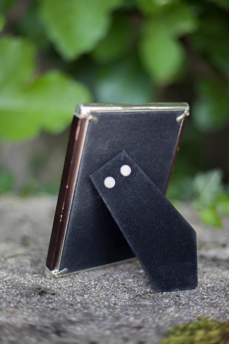 May include: A small, rectangular picture frame with a dark brown border and a silver-colored trim. The back of the frame is black, with a black velvet easel stand. The frame is set on a gray stone surface, with green foliage in the background.