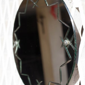 May include: A round, black framed mirror with a starburst design etched into the glass. The mirror has a green tint and is hanging on a white wall.
