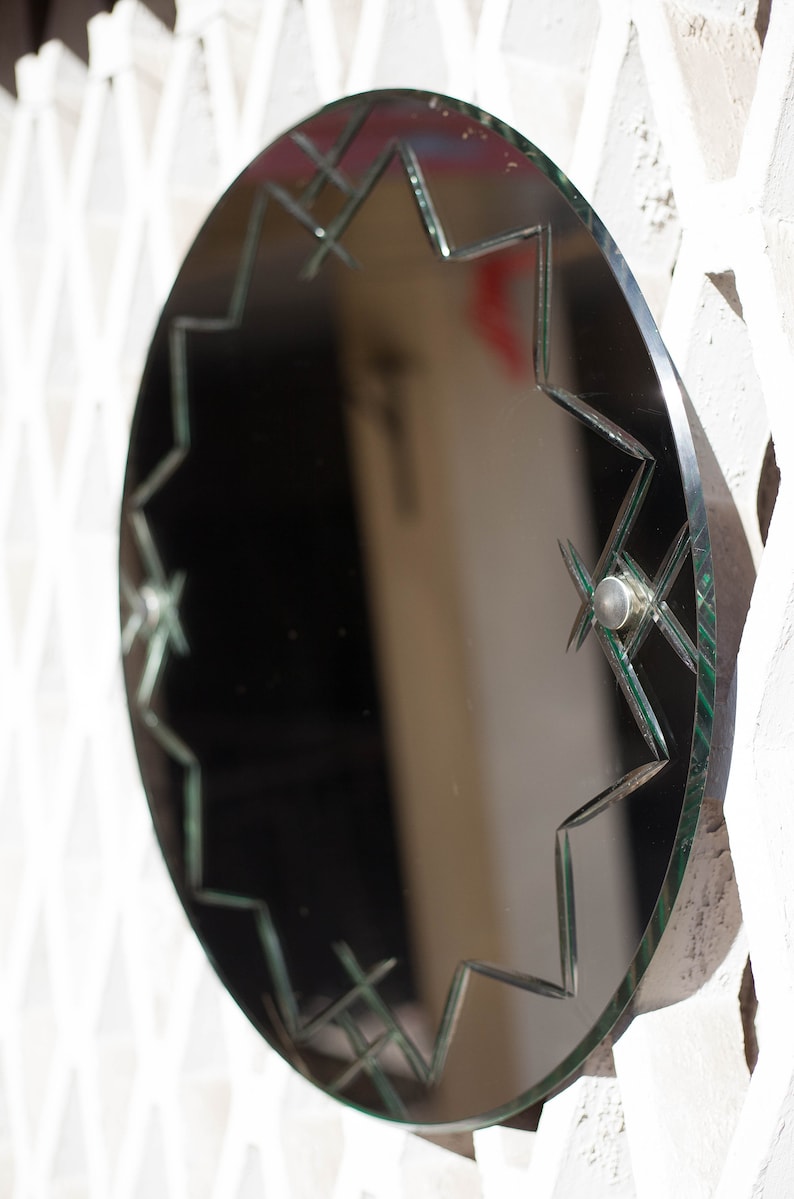 May include: A round, black framed mirror with a starburst design etched into the glass. The mirror has a green tint and is hanging on a white wall.