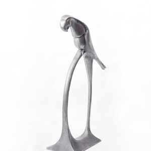 Op de afbeelding: Een zilveren metalen sculptuur van een papegaai die op een gebogen basis staat. De papegaai zit in een gehurkte positie met zijn hoofd naar beneden gekanteld.