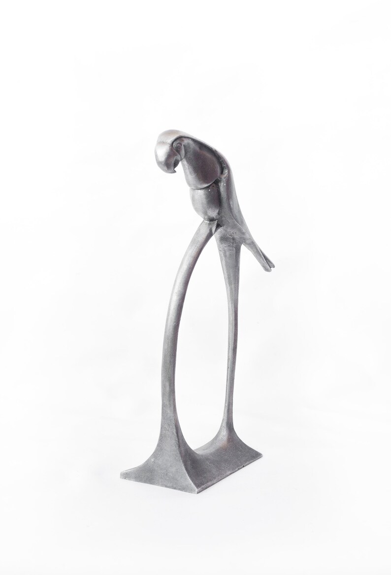 Op de afbeelding: Een zilveren metalen sculptuur van een papegaai die op een gebogen basis staat. De papegaai zit in een gehurkte positie met zijn hoofd naar beneden gekanteld.
