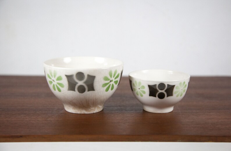 May include: Two white ceramic bowls with a geometric pattern of green and black flowers. The bowls are on a wooden surface.
