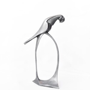 Op de afbeelding: Een zilveren metalen sculptuur van een papegaai die op een gebogen basis staat. De papegaai kijkt naar rechts en heeft zijn kop naar beneden gekanteld.