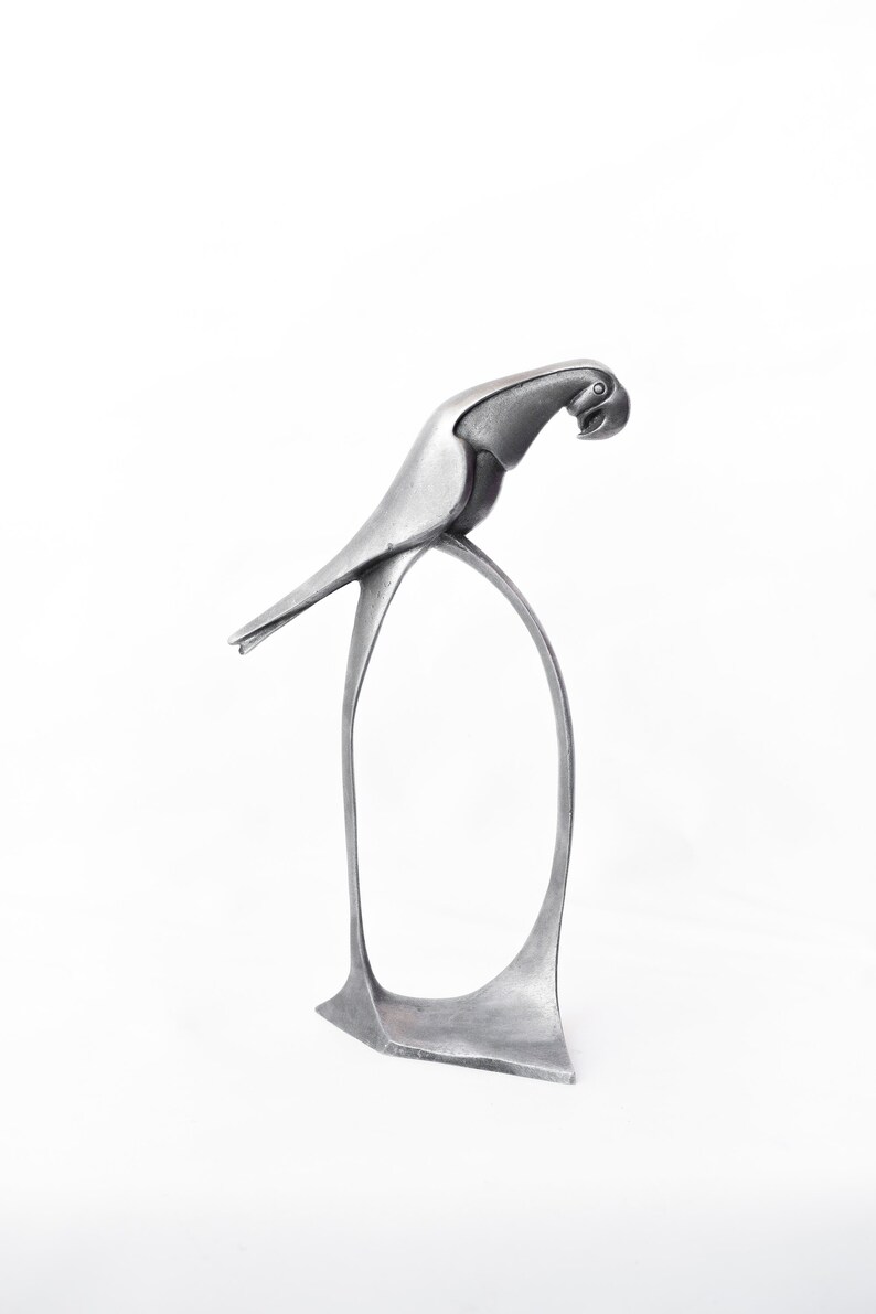 Op de afbeelding: Een zilveren metalen sculptuur van een papegaai die op een gebogen basis staat. De papegaai kijkt naar rechts en heeft zijn kop naar beneden gekanteld.