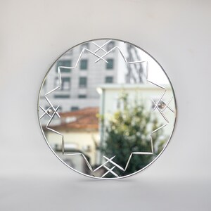 May include: A round, silver-framed mirror with a geometric design etched into the glass. The mirror reflects a blurry image of a building and trees.