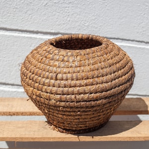 Peut inclure: Un grand panier rond en osier brun, fait de matériaux naturels. Le panier est posé sur une surface en bois.