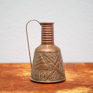 Può includere: Un piccolo brocca di rame con un design decorato. La brocca ha un manico e una base larga.