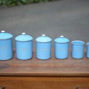 May include: Set of six blue enamel canisters with white lids and white lettering. The canisters are labeled "Sugar", "Coffee", "Tea Bags", "Flour", "Spice", and "Rice".