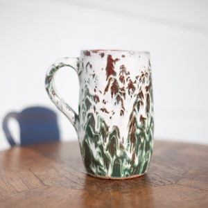 May include: A white ceramic mug with a green and brown marbled glaze. The mug has a rounded body and a handle.