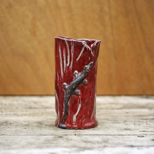 Può includere: Un vaso in ceramica rossa con una superficie strutturata e un design di lucertola nera. Il vaso è appoggiato su una superficie di legno.