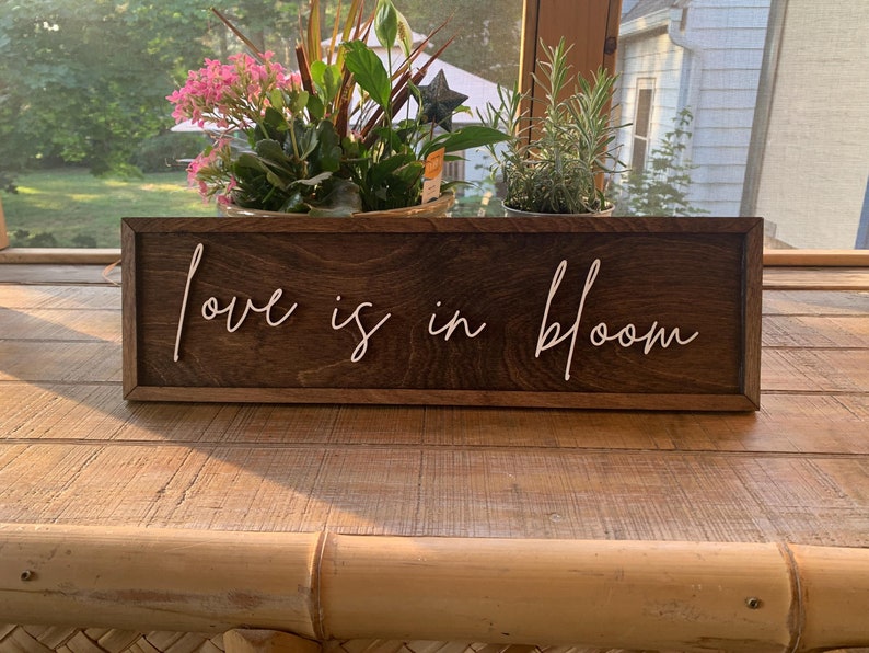 Op de afbeelding: Een houten bord met de tekst "Love is in Bloom" geschreven in witte, cursieve letters. Het bord staat op een houten tafel.