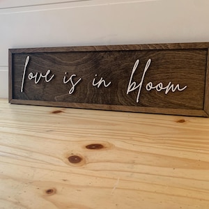 Op de afbeelding: Een houten bord met de tekst "Love is in bloom" geschreven in witte, cursieve letters.