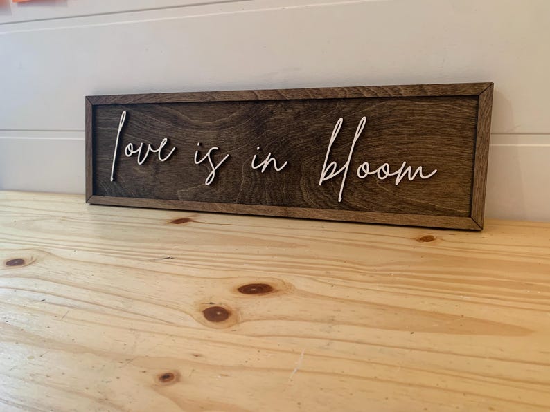 Op de afbeelding: Een houten bord met de tekst "Love is in bloom" geschreven in witte, cursieve letters.