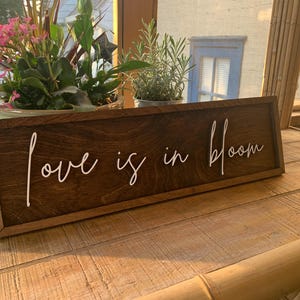 Op de afbeelding: Een houten bord met een bruine vlek en een wit geschilderd opschrift dat "love is in bloom" luidt.