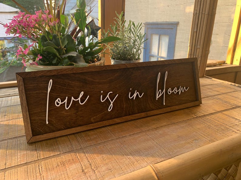 Op de afbeelding: Een houten bord met een bruine vlek en een wit geschilderd opschrift dat "love is in bloom" luidt.