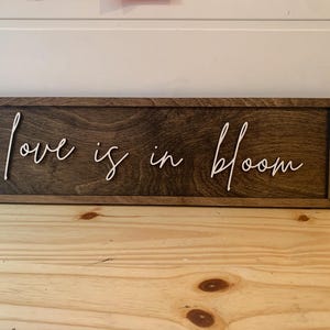Op de afbeelding: Een houten bord met de tekst "Love is in bloom" geschreven in witte, cursieve letters.