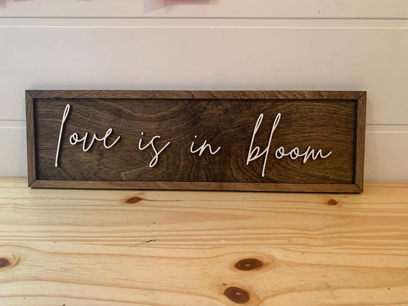 Op de afbeelding: Een houten bord met de tekst "Love is in bloom" geschreven in witte, cursieve letters.