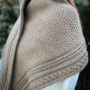 May include: A close-up of a beige knitted shawl. The shawl has a textured pattern with a cable knit border. The garment is draped over the shoulders, showcasing the intricate stitch work and warm color.