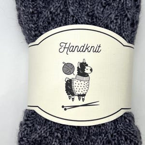 以下が含まれることがあります： 装飾的な模様の、濃い灰色の手編みの靴下。 「Handknit」の文字と、編み物をしているラマのイラストが描かれたクリーム色のラベルが靴下を包んでいます。 銀色のボタンが付いた茶色の革製のタグが取り付けられています。