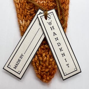May include: Close-up of a hand-knitted item in shades of orange and brown. Two rectangular tags are attached with a brown string. One tag reads "MADE BY" and the other says "HAND KNIT" in black letters.
