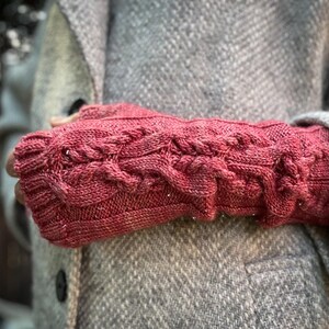 Puede incluir: Guantes sin dedos rojos con diseño de punto trenzado. Los guantes están hechos de un hilo texturizado y se usan sobre un abrigo gris. Un accesorio cálido para el clima frío.