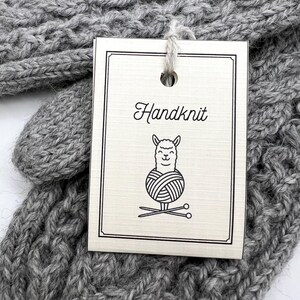 Op de afbeelding: Close-up van een grijs handgebreid item met een label. Het label leest "Handknit" met een illustratie van een lama die een bol wol en breinaalden vasthoudt. Het gebreide item heeft een textuurpatroon en is gemaakt van zacht, grijs garen.