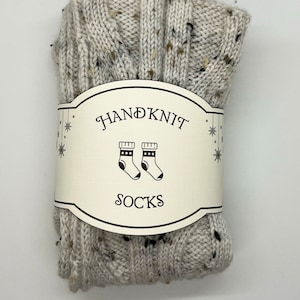 Puede incluir: Un par de calcetines tejidos a mano de color beige moteado con una etiqueta blanca que dice "HAND KNIT SOCKS" y muestra un par de calcetines con rayas blancas y negras.