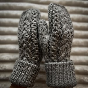 May include: A pair of gray knitted mittens with a cable knit design. The mittens have ribbed cuffs and a textured pattern throughout the body. The background is a soft, neutral color.