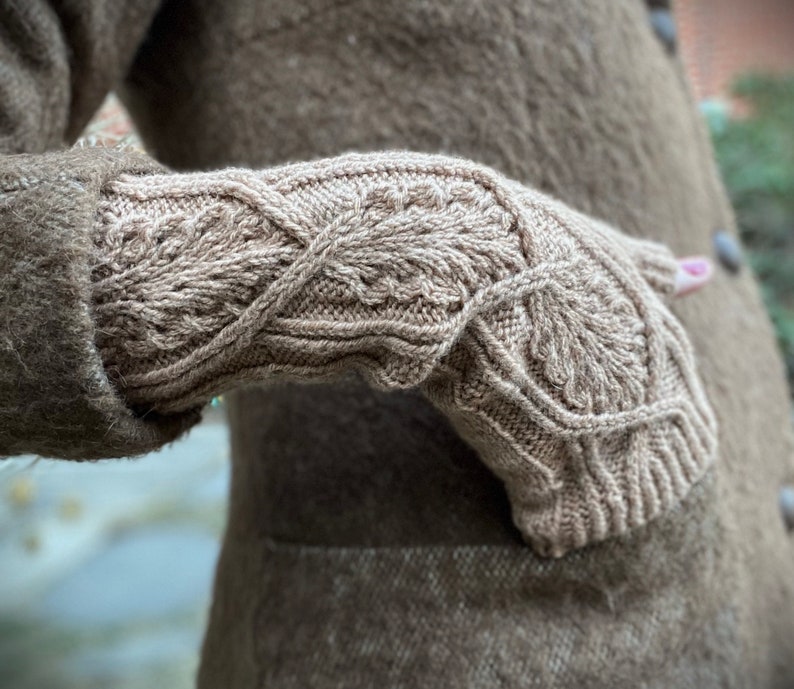 May include: A close-up of a brown knitted mitten with a detailed cable knit pattern. The mitten is worn by a person wearing a brown coat.