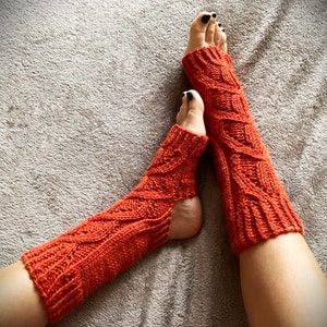 Pode incluir: Um par de polainas de malha laranja com um padrão de malha de cabo. As polainas são usadas nas pernas de uma pessoa, com os pés a sair da parte inferior.