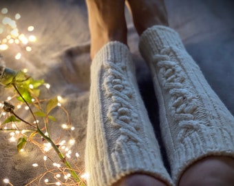 May include: A pair of white knitted leg warmers with a cable knit pattern. The leg warmers are worn by a person and are on a light brown surface with string lights.