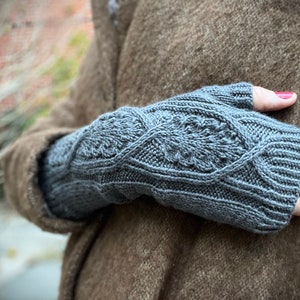 May include: Gray fingerless gloves with a cable knit pattern. The gloves are worn by a person wearing a brown coat.