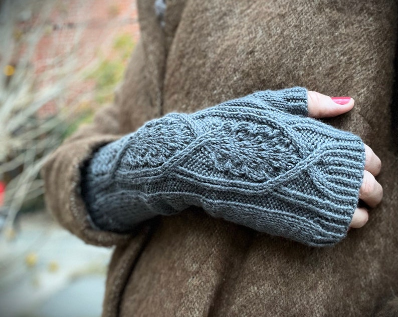 May include: Gray fingerless gloves with a cable knit pattern. The gloves are worn by a person wearing a brown coat.