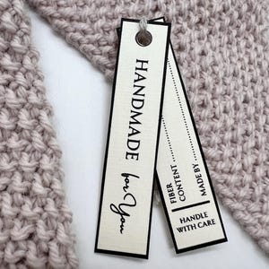 Op de afbeelding: Close-up van een gebreid item in een zachte, gedempte mauve kleur. Twee rechthoekige labels zijn bevestigd met een grijze draad. De labels lezen "HANDMADE for You", "FIBER CONTENT" en "MADE BY HANDLE WITH CARE".