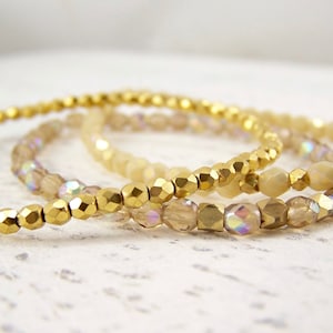 May include: Three gold bracelets with iridescent glass beads. The bracelets are stacked on top of each other.