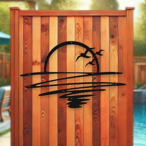 Puede incluir: Silueta de metal negro de una puesta de sol sobre el agua con pájaros volando por encima. La silueta está montada en una valla de madera.