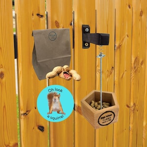 May include: A wooden squirrel feeder with a hexagonal shape and a metal spring-loaded mechanism. The feeder is mounted on a wooden fence and is filled with peanuts. A blue and white sticker with the text "Oh look a squirrel!" is attached to the feeder. A brown paper bag with the text "Duck & Green" is hanging on the fence.