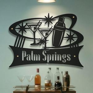 May include: Black metal wall art featuring a cocktail shaker, martini glasses, and starburst designs. The words "Palm Springs" are displayed in a banner below the design. A bar with bottles and glasses is visible below.