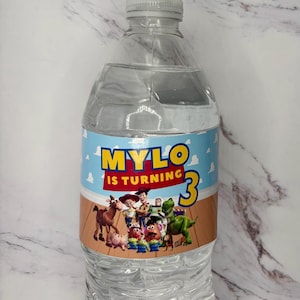 May include: A clear plastic water bottle with a custom label featuring a light blue sky and wood floor background. The text reads MYLO IS TURNING 3 in yellow and red letters. The label includes a group of animated toy characters from Toy Story.