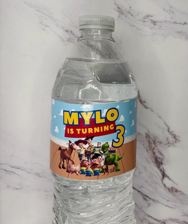 May include: A clear plastic water bottle with a custom label featuring a light blue sky and wood floor background. The text reads MYLO IS TURNING 3 in yellow and red letters. The label includes a group of animated toy characters from Toy Story.
