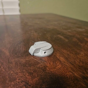 Puede incluir: Un objeto de metal plateado con una tapa con bisagras y un pequeño agujero en el centro. El objeto está sobre una superficie de madera.