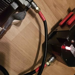 May include: Close-up of a black device with a red light and a silver connector. A black corrugated hose with red tape is connected to the device. Two red cylindrical objects are on the device.