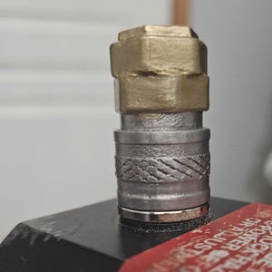 May include: Close-up of a metal fitting with a gold-colored top section and a silver-colored lower section. The lower section has a textured pattern. The fitting is attached to a black surface, with a red label visible.