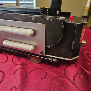 May include: A black and silver toy train with a red button and two silver panels with raised details. The train has a worn, distressed look, giving it a vintage feel.