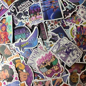 May include: A collection of colorful stickers featuring anime-style illustrations and text. The stickers showcase various designs, including characters, logos, and phrases like "Demon Hunters" and "K-Pop." The color palette includes purple, pink, and blue.